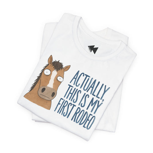 A folded white t-shirt featuring a cartoon illustration of a brown horse with a white blaze and the text 'ACTUALLY, THIS IS MY FIRST RODEO' printed in blue. A small black lightning bolt logo is visible inside the collar.