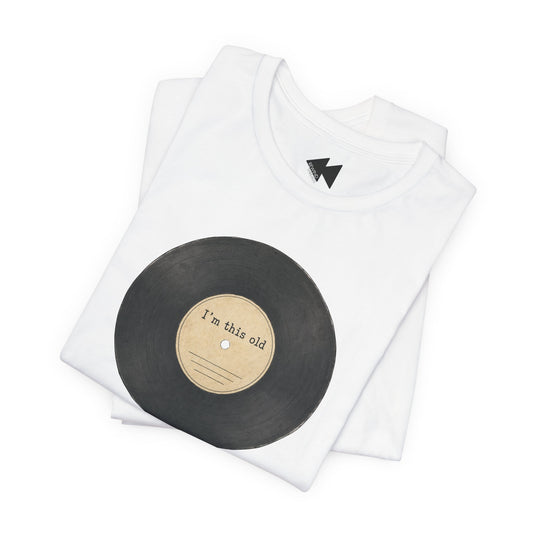 A folded white t-shirt featuring a graphic of a black vinyl record with a beige center label that reads 'I'm this old'.