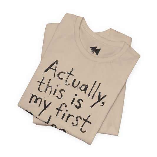 A folded beige t-shirt with black handwritten-style text reading 'Actually, this is my first...' on a plain white background.