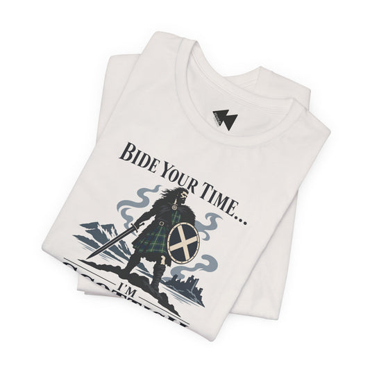 A folded white t-shirt with a graphic print of a Scottish warrior in a tartan kilt holding a sword and shield, with the text 'BIDE YOUR TIME.