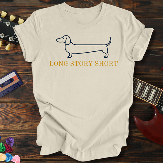 Beige t-shirt with dachshund graphic and 'Long Story Short' text on a wooden surface with guitar and cassette tape.