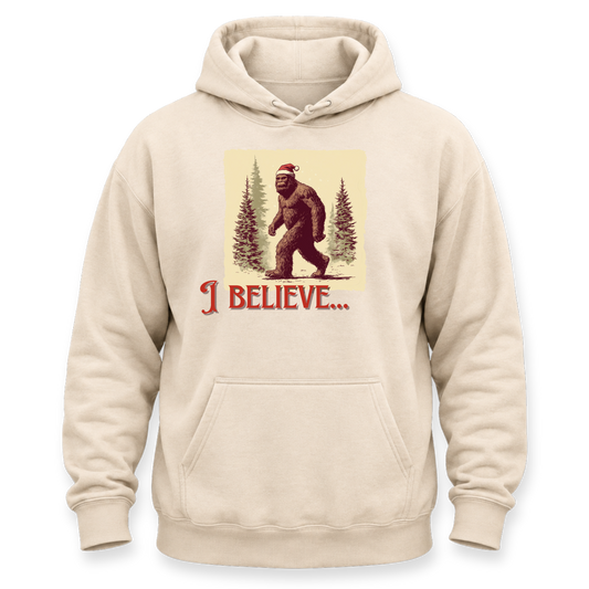 A beige pullover hoodie with a front graphic of a Bigfoot figure wearing a Santa hat among pine trees, with the text 'I BELIEVE.