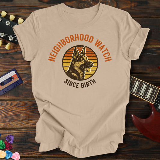 Beige t-shirt with a dog graphic and text 'Neighborhood Watch Since Birth' on a wooden surface with a guitar and cassette tape.