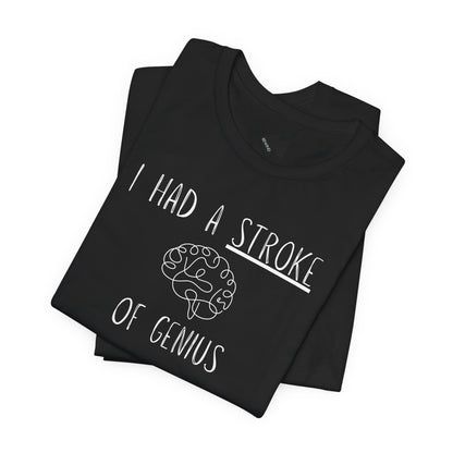 A folded black t-shirt with a black graphic print that reads 'I HAD A STROKE OF GENIUS' surrounding a simple line drawing of a brain. A small black geometric logo is visible inside the collar.