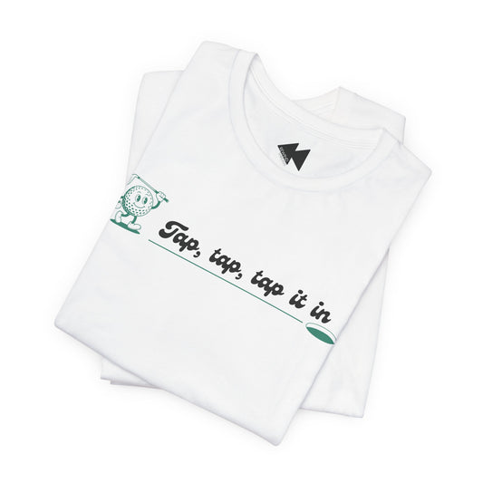 A folded white t-shirt with a green and black graphic on the chest that reads 'Tap, tap, tap it in' next to a cartoon character, shown on a clean white background.