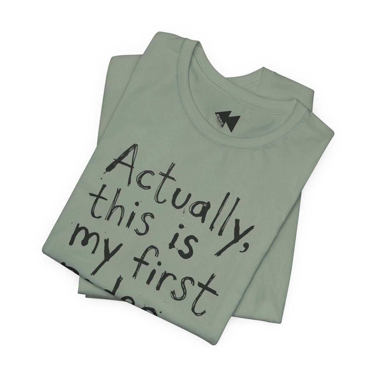 A folded sage t-shirt with black handwritten-style text reading 'Actually, this is my first...' on a plain white background.