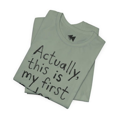 A folded sage t-shirt with black handwritten-style text reading 'Actually, this is my first...' on a plain white background.