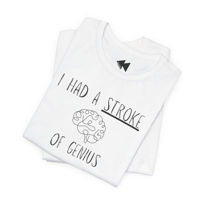 A folded white t-shirt with a black graphic print that reads 'I HAD A STROKE OF GENIUS' surrounding a simple line drawing of a brain. A small black geometric logo is visible inside the collar.