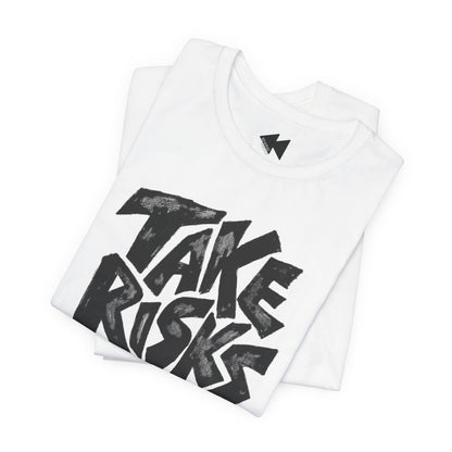 A folded white t-shirt with a large black graphic print reading 'TAKE RISKS' in a bold, hand-drawn style. A small black lightning bolt logo is visible inside the collar.