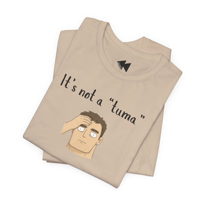 A folded beige t-shirt with a black graphic print that reads 'It's not a "tuma"' above an illustration of a man scratching his head. The shirt features a small black geometric logo printed inside the collar.