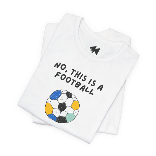 A folded white t-shirt with a graphic print that reads 'NO, THIS IS A FOOTBALL' above a hand-drawn style soccer ball in black, blue, yellow, and mint green. A small black geometric logo is printed inside the collar.