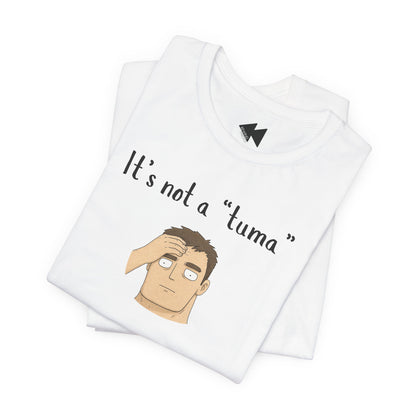 A folded white t-shirt with a graphic print that reads 'It's not a "tuma"' above an illustration of a man with brown hair covering his face. The shirt features a black geometric logo inside the collar.