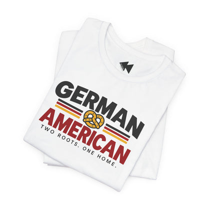 A folded white t-shirt with a graphic print that reads 'GERMAN AMERICAN' in black and red lettering, featuring a yellow pretzel symbol and the tagline 'TWO ROOTS. ONE HOME.