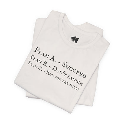 A folded light beige short-sleeve crewneck t-shirt with black printed text on the front reading 'PLAN A. - SUCCEED PLAN B. - DON'T PANICK PLAN C. RUN FOR THE HILLS'