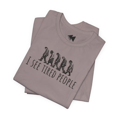 Stack of folded dusty pink short-sleeve t-shirts with a black graphic of five progressively slacker-looking people walking side-by-side above the text 'I SEE TIRED PEOPLE'.