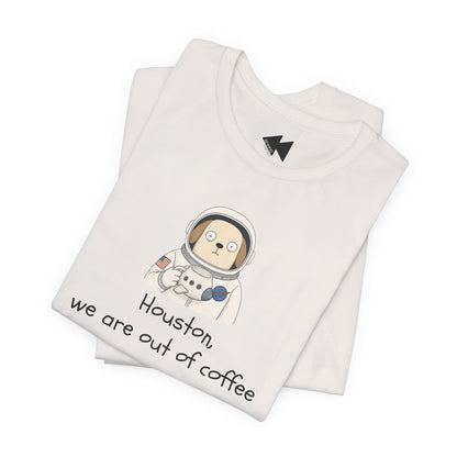 A folded light beige t-shirt with a cartoon astronaut dog graphic and the text 'Houston, we are out of coffee' printed on the front, displayed on a clean white background.
