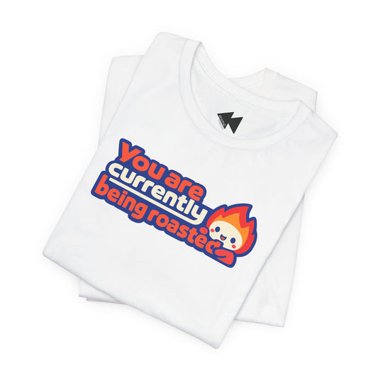 A folded white t-shirt with a graphic print that reads 'You are currently being roasted' in red and blue bubble letters, next to a cartoon fire character, on a clean white background.