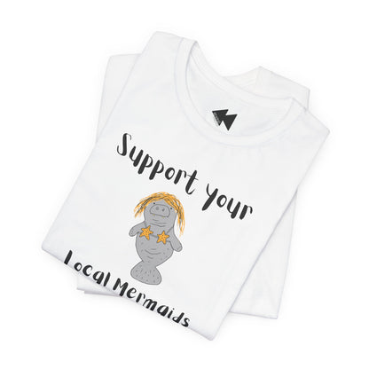 A folded white t-shirt with a graphic of a gray mermaid wearing a yellow pineapple headband and star necklace, with the text 'Support your Local Mermaids' in black script.