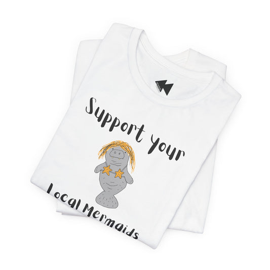A folded white t-shirt with a graphic of a gray mermaid wearing a yellow pineapple headband and star necklace, with the text 'Support your Local Mermaids' in black script.