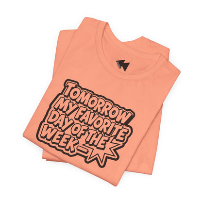 A folded peach-colored t-shirt with a bold graphic reading 'TOMORROW MY FAVORITE DAY OF THE WEEK' in black and white bubble letters, set against a plain white background.