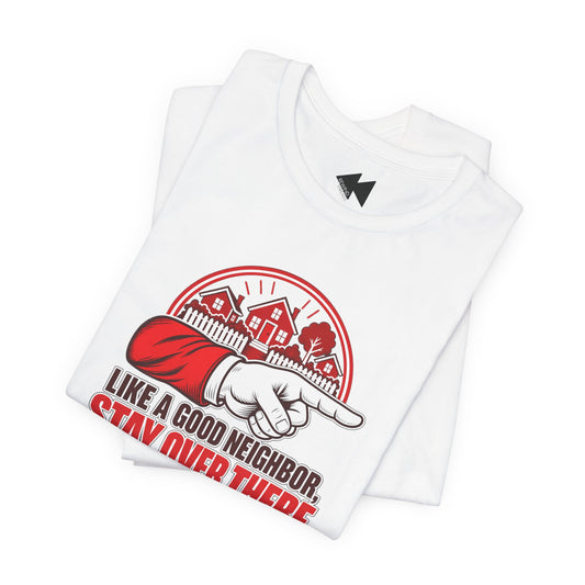 A folded white t-shirt with a graphic print on the front. The graphic features a red and black circular emblem with houses, a pointing hand in a red sleeve, and the text 'LIKE A GOOD NEIGHBOR STAY OVER THERE'.