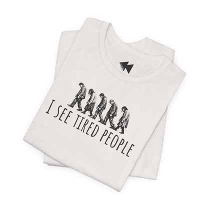 A folded white t-shirt with a black graphic print showing the evolution of humans walking and the text 'I SEE TIRED PEOPLE'. A small black geometric logo is printed inside the collar.