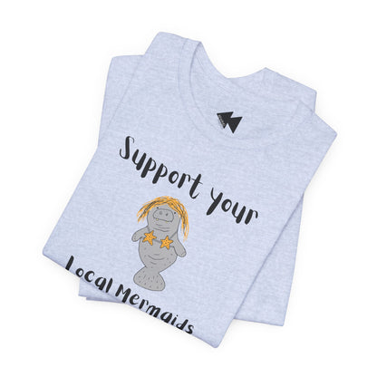 A light heather gray folded t-shirt featuring a cartoon manatee graphic with yellow hair and starfish, accompanied by the text 'Support your Local Mermaids' in black script.