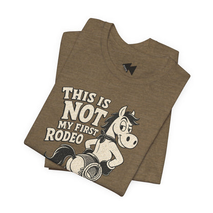 A folded brown heather t-shirt featuring a graphic print of a cartoon horse wearing a cowboy hat and bandana, with the text 'THIS IS NOT MY FIRST RODEO' in a distressed cream font.