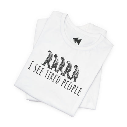 A folded white t-shirt with a black graphic print showing a line of walking figures and the text 'I SEE TIRED PEOPLE'. A small black lightning bolt logo is printed inside the collar.