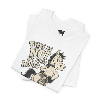 A folded white t-shirt featuring a graphic print of a cowboy riding a bucking horse with the text 'THIS IS NOT MY FIRST RODEO' in a distressed beige font. The shirt is displayed on a clean white background.