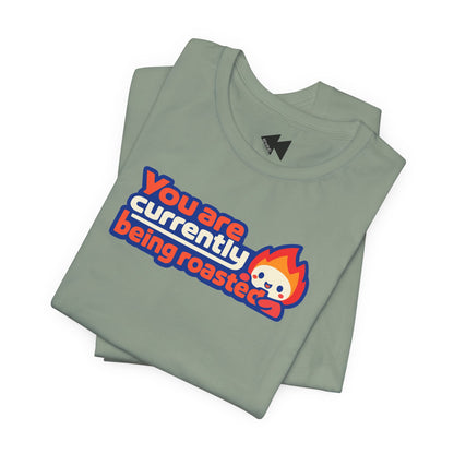 A folded sage green t-shirt with a graphic that reads 'You are currently being roasted' in red, white, and blue, next to a cartoon fire character, on a white background.