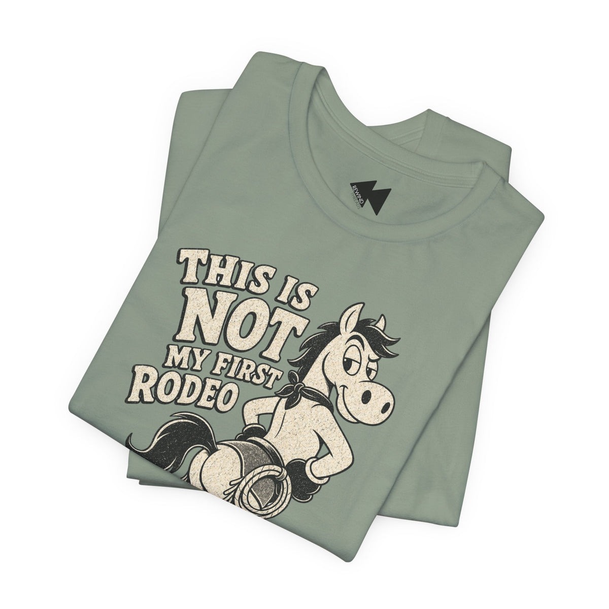 A folded sage green t-shirt featuring a graphic of a cowboy riding a bucking horse with the text 'THIS IS NOT MY FIRST RODEO' in a distressed cream font, set against a plain white background.