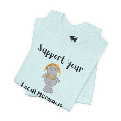 A folded light blue heather t-shirt featuring a cartoon illustration of a mermaid manatee with yellow hair and starfish, accompanied by the text 'Support your Local Mermaids' in black script.
