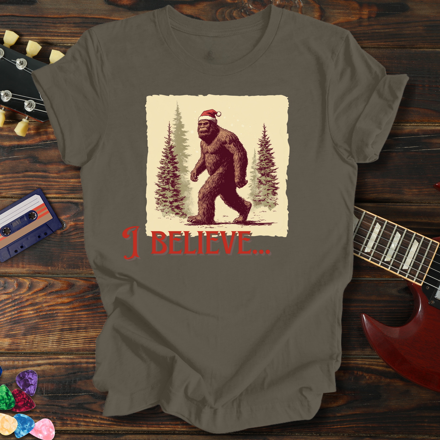 Gray t-shirt with a graphic of a bear wearing a Santa hat and 'I Believe' text on a wooden surface with a guitar and cassette tape.