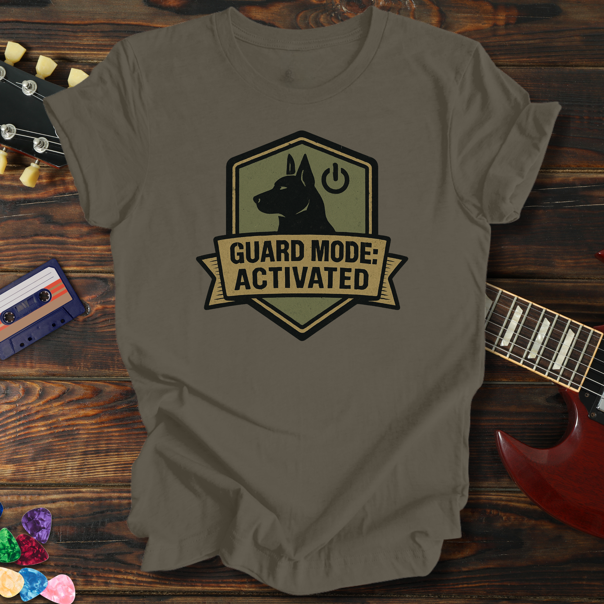 Brown t-shirt with 'Guard Mode: Activated' graphic on a wooden surface with a guitar and cassette tape.