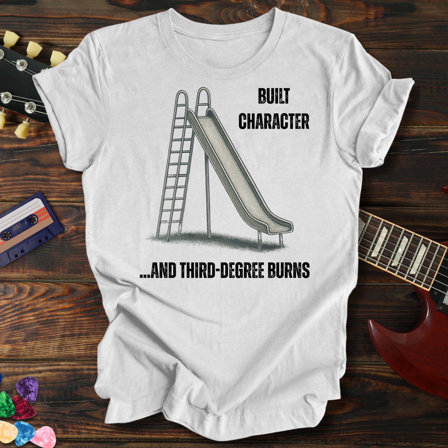White t-shirt with a graphic of a slide and ladder, text 'BUILT CHARACTER AND THIRD-DEGREE BURNS', on a wooden surface with a guitar and cassette tape.