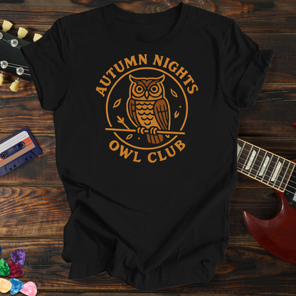 Black t-shirt with 'Autumn Nights Owl Club' design on a wooden surface with a guitar and cassette tape.