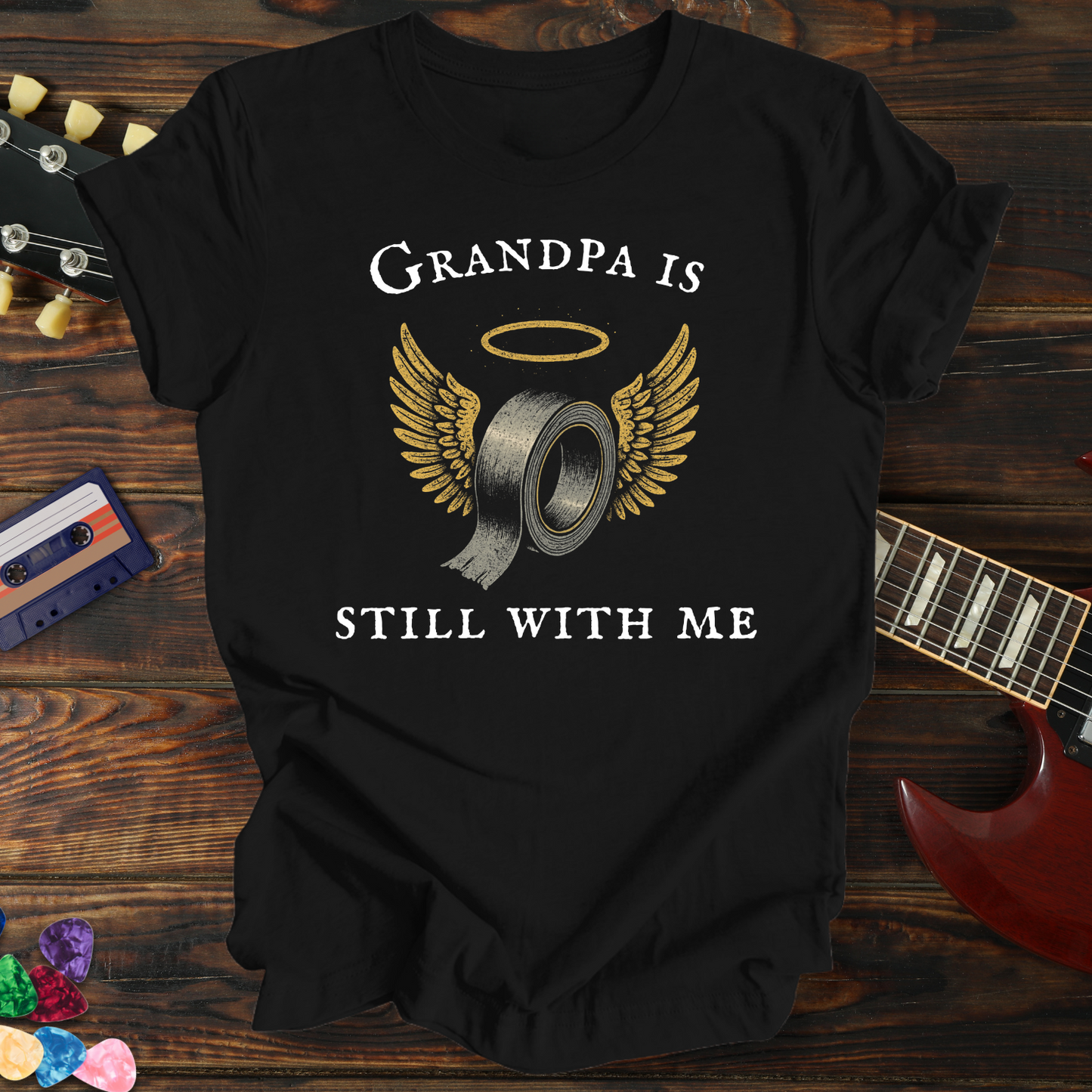 Black t-shirt with 'Grandpa is still with me' design on a wooden surface with a guitar and cassette tape.