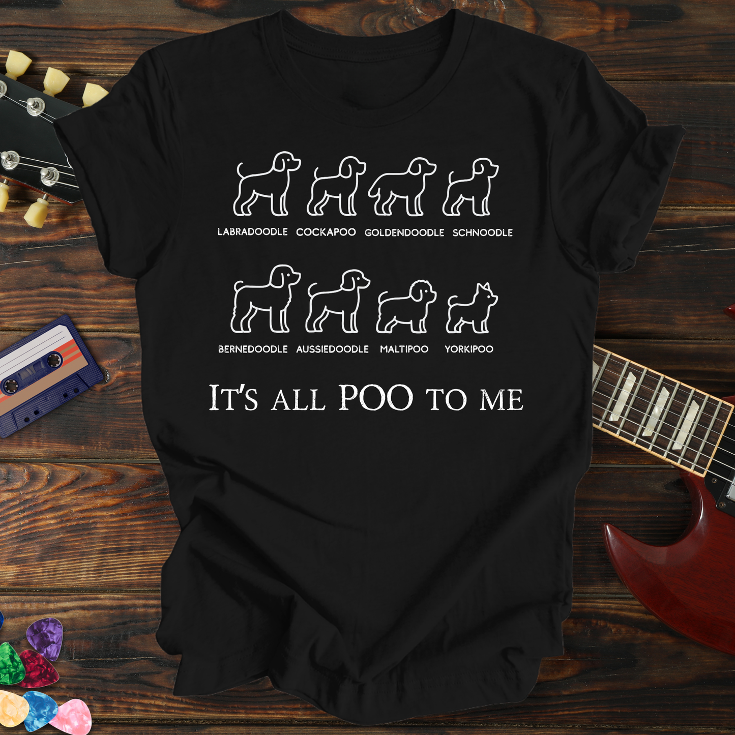 Black t-shirt with dog breed illustrations and text on a wooden surface with a guitar and cassette tape.
