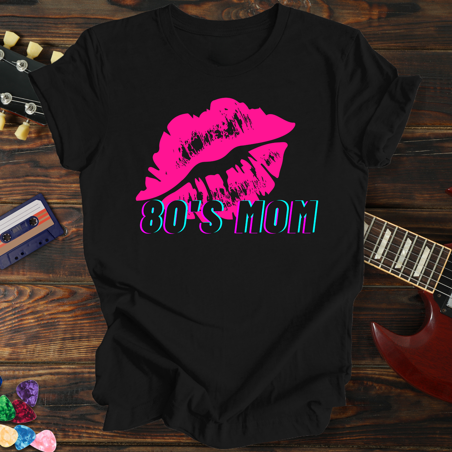 Black t-shirt with pink lips and '80's Mom' text on a wooden surface with a guitar and cassette tape.