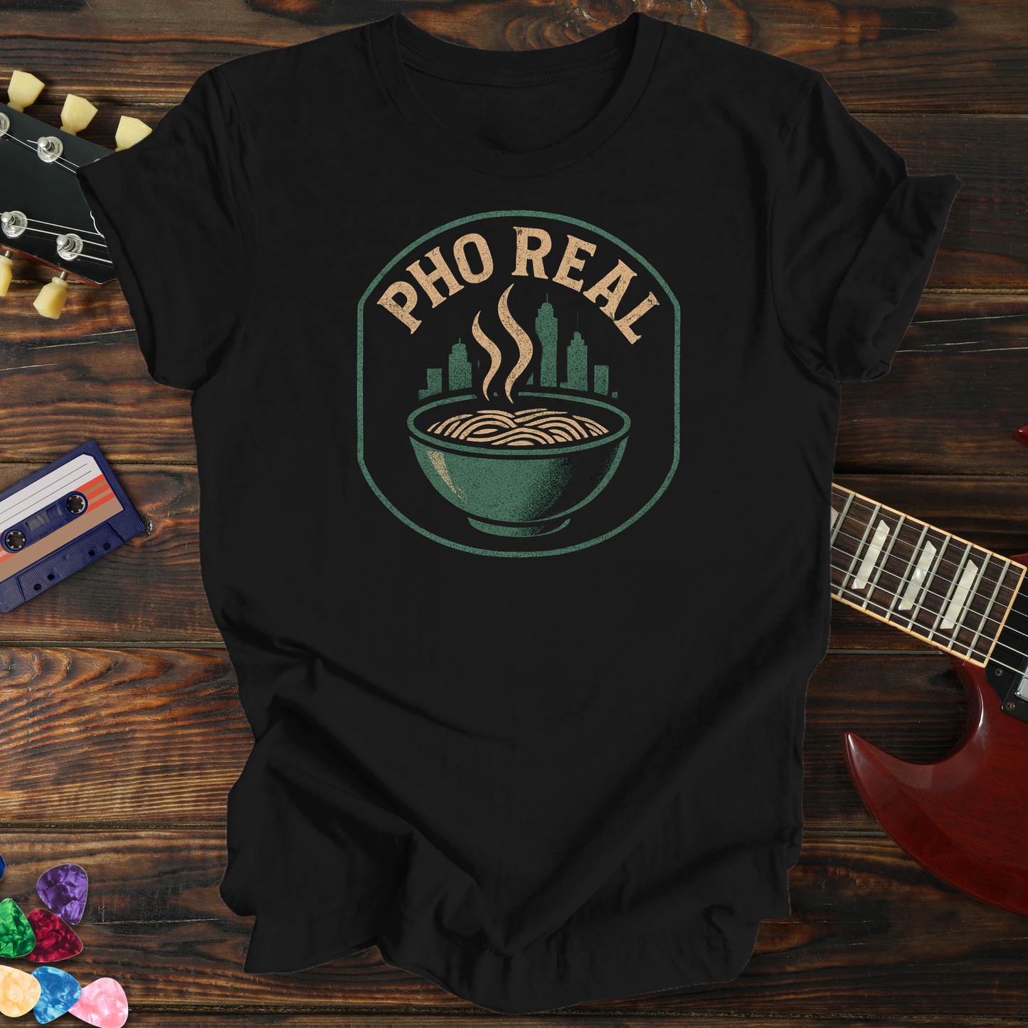 Black t-shirt with 'Pho Real' graphic design on a wooden surface with a guitar and cassette tape.