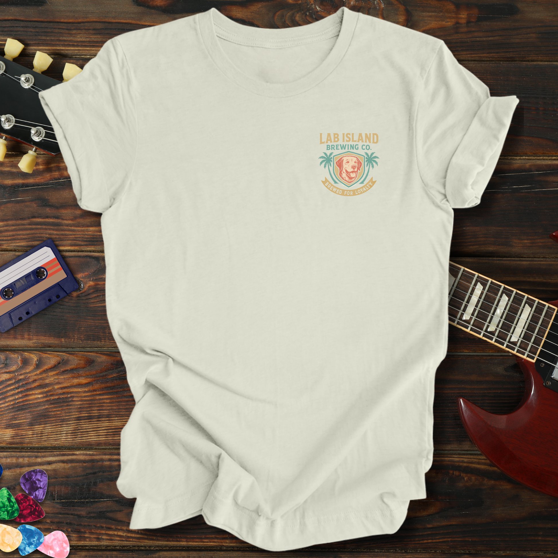White t-shirt with a logo on a wooden surface with a guitar and cassette tape.