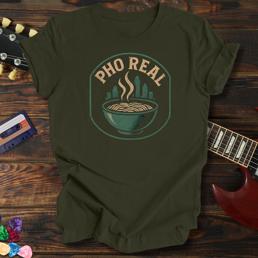 Green t-shirt with 'Pho Real' graphic on a wooden surface with a guitar and cassette tape.