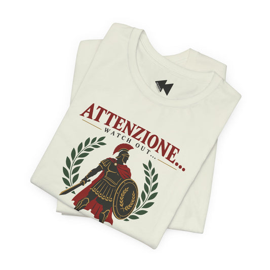 A folded cream-colored t-shirt featuring a graphic print of a Roman gladiator with a shield and helmet, flanked by laurel wreaths, with the Italian text 'ATTENZIONE.