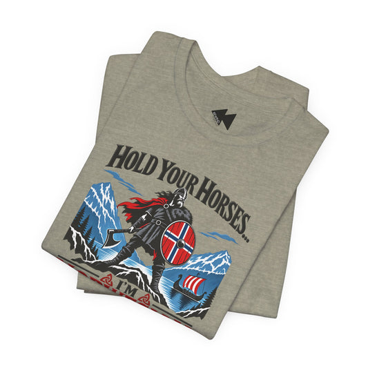 A folded heather gray t-shirt featuring a graphic print of a Viking warrior with a red and blue shield, mountains, and the text 'HOLD YOUR HORSES.