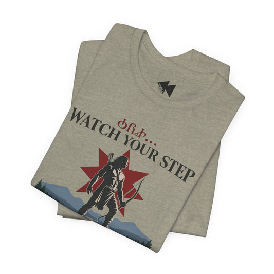 A folded heather gray t-shirt featuring a graphic print of a black silhouette archer standing in front of a red star, with the text 'WATCH YOUR STEP' in black and red Greek letters above it. The shirt is displayed on a clean white background.