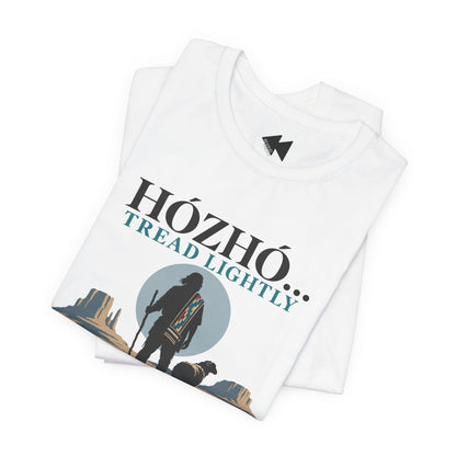 A folded white t-shirt with a graphic print on the front. The graphic features the text 'HÓZHÓ TREAD LIGHTLY' in black and teal lettering, with a silhouette of a hiker carrying a patterned backpack against a circular blue-gray background and desert rock formations at the bottom.