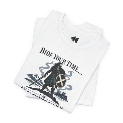 A folded white t-shirt with a graphic print of a Scottish warrior in a tartan kilt holding a sword and shield, with the text 'BIDE YOUR TIME.