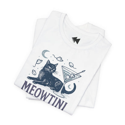 A folded white t-shirt featuring a dark navy blue graphic of a cat sitting next to a martini glass with olives, surrounded by small mouse illustrations, with the word 'MEOWTINI' printed below. A small black lightning bolt logo is visible inside the collar.