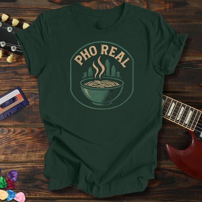 Green t-shirt with 'Pho Real' design on a wooden surface with a guitar and cassette tape.